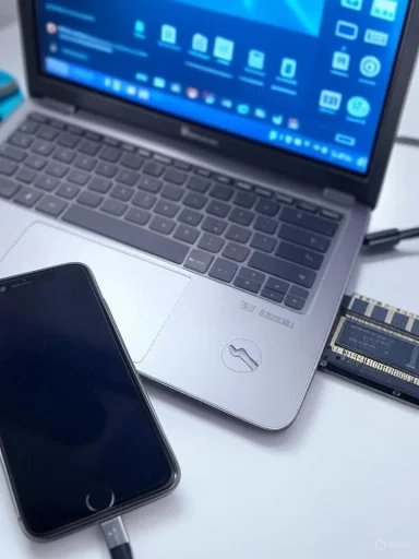 A laptop, smartphone, and solid-state drive on a white surface, with the laptop screen displaying a blue desktop background.